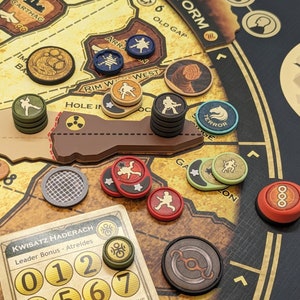 Puede incluir: Un juego de mesa con varias piezas esparcidas sobre un tablero de juego marrón. Las piezas incluyen fichas circulares en colores como rojo, verde y azul, y una tarjeta con el texto "KWISATZ HADERACH". El tablero tiene un diseño de mapa.