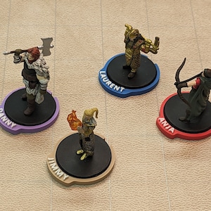 Puede incluir: Cuatro figuras en miniatura sobre bases negras con anillos de colores alrededor de los bordes. Los anillos están etiquetados como "Danny", "Jimmy", "Laurent" y "Anja".