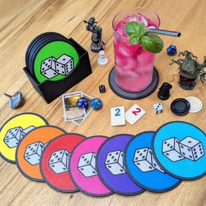 May include: A selection of colourful dice-themed coasters, a drink, and game pieces on a wooden surface. The coasters are in various colours, each with a pair of dice. Other items include dice, game cards, and miniature figurines.