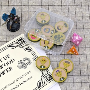 May include: A collection of tabletop gaming accessories, including character tokens, dice, and a miniature figure. The tokens are round with goblin-like character designs and numbers. A clear plastic container holds some of the tokens. A booklet is also present.