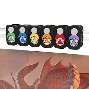 Op de afbeelding: Een set van zes zwarte rechthoekige clips, elk met een witte ronde bovenkant en een gekleurd basisgedeelte in groen, blauw, geel, oranje, rood en paars. Elke clip toont een afbeelding van een witte twintigzijdige dobbelsteen met een kleur.
