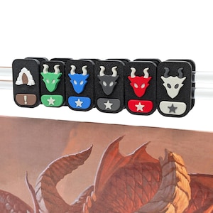 Puede incluir: Un conjunto de seis clips rectangulares negros sujetos a una varilla transparente, cada uno con un icono de cabeza de dragón en verde, azul, gris oscuro, rojo o blanco, junto a una base de color que muestra símbolos como estrellas o un signo de exclamación.