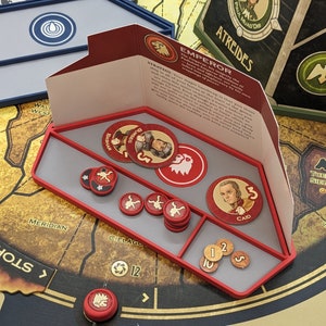 Puede incluir: Una bandeja de juego de plástico rojo con compartimentos para guardar las piezas del juego. La bandeja tiene un interior gris y está etiquetada con las palabras "Emperador", "Atreides", "Caid", "Cielage", "Meridian" y "Tormenta".