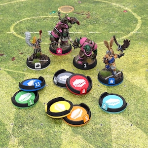 Puede incluir: Figuras y fichas de fútbol de fantasía en miniatura dispuestas sobre un campo verde. Las figuras incluyen orcos y un personaje parecido a un conejo. Las fichas son negras con centros de colores y gráficos de fútbol, ​​numeradas del 1 al 16.