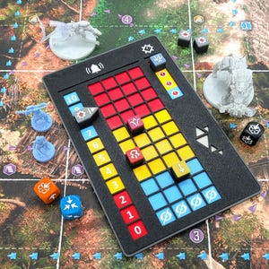 May include: A black rectangular board game dashboard with a grid of red, yellow, and blue squares, numbered zero to ten. It features small coloured cubes, a bell icon, and various game pieces including miniature figures and dice on a printed map.