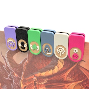 Puede incluir: Seis clips magnéticos rectangulares en morado, negro, verde, azul, blanco y rosa. Cada clip presenta una incrustación circular beige con un icono único en relieve blanco o rosa, incluyendo un ojo, una calavera, una hoja, una capucha, un engranaje y un símbolo de instrumento musical.