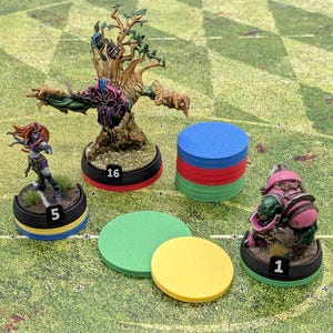 Puede incluir: Tres miniaturas de juegos de mesa pintadas y una pila de fichas de plástico de colores. Las figuras incluyen una criatura arbórea, un humanoide de pelo naranja y un personaje corpulento con armadura, cada uno sobre una base numerada: 16, 5 y 1.