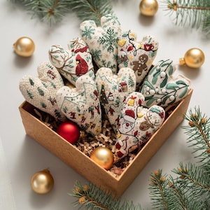 May include: A collection of heart-shaped fabric ornaments featuring intricate embroidery of Christmas themes including a cardinal, gingerbread man, Santa Claus, reindeer, mistletoe, and winter landscapes, nestled in a brown cardboard box.