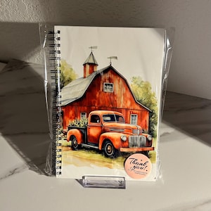 May include: Spiral-bound notebook featuring a watercolor illustration of a red barn and an orange vintage pickup truck. The artwork has a rustic, country theme. A small round sticker with the words "Thank you!" is visible.