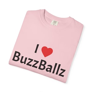 May include: A light pink t-shirt with the text "I ❤️ BuzzBallz" in black and a red heart. The shirt is folded and appears to be made of a soft material. The tag is visible in the upper right corner.