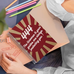 May include: Spiral-bound notebook with a burgundy cover featuring the number "444" and the text "The Universe & your guides are always protecting you!" in white. The design includes gold sunburst rays. Pencils and paperclips are also visible.