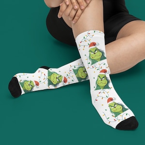 May include: White crew socks featuring a repeating pattern of a green, frowning character wearing a red and white Santa hat, surrounded by colorful, tangled Christmas light strings. The socks have solid black toe and heel sections.