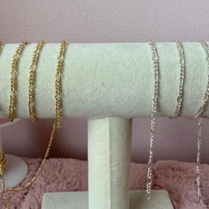 May include: Gold and silver Figaro chain necklaces displayed on a white stand. The gold chains are on the left, and the silver chains are on the right. The chains are draped over a cylindrical display.