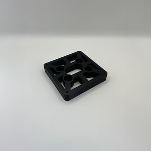 May include: A black, square-shaped object with a grid-like design. The object has four circular holes and a series of rectangular cutouts. The item is made of a solid, matte black material and sits on a white surface.