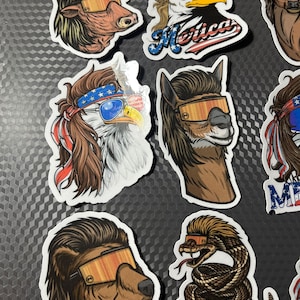 May include: A set of nine colorful stickers featuring various animals wearing sunglasses and bandanas. The animals include a bull, an eagle, an alpaca, a snake, a bear, and a deer. The stickers have a patriotic theme with red, white, and blue colors and the word "Merica" on one of the stickers.
