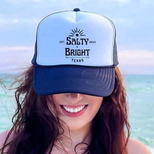 May include: A woman wearing a white and black trucker hat with the text "EST. 2023 SALTY & BRIGHT TEXAS" on the front. She is smiling and has long brown hair.
