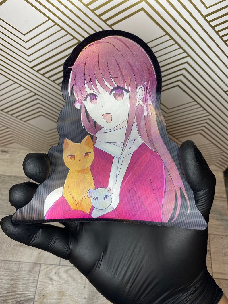 Puede incluir: Una pegatina hologr&aacute;fica con un personaje de anime de pelo largo rosa, que lleva un su&eacute;ter rosa y blanco, sosteniendo un gato amarillo y un rat&oacute;n blanco.