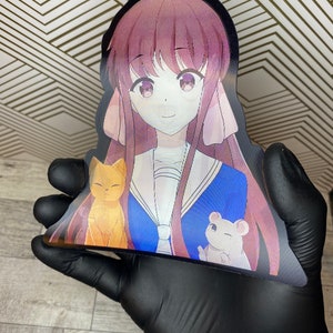 Puede incluir: Una pegatina con una chica de anime de dibujos animados con el pelo largo de color rosa que lleva un uniforme escolar azul y blanco. Est&aacute; sosteniendo un gato amarillo y un rat&oacute;n blanco.