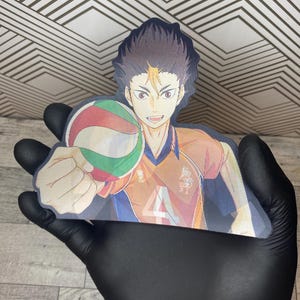 Puede incluir: Una pegatina con un personaje de anime con cabello casta&ntilde;o y ropa naranja y azul sosteniendo un bal&oacute;n de voleibol. El personaje sonr&iacute;e y tiene una expresi&oacute;n decidida. La pegatina est&aacute; impresa sobre un fondo blanco.