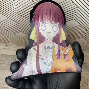 Puede incluir: Un recorte de un personaje de anime con cabello largo morado y cintas amarillas. El personaje lleva un uniforme de marinero blanco con un cuello azul y una corbata de lazo roja. El personaje sonr&iacute;e y tiene ojos grandes y blancos. El personaje sostiene un peque&ntilde;o oso marr&oacute;n y un gato naranja grande con una melena amarilla. El gato est&aacute; rugiendo.