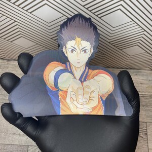 Puede incluir: Un personaje de anime en blanco y negro con ropa naranja y azul se representa en un primer plano. El personaje est&aacute; extendiendo su mano derecha, con los dedos extendidos, como para agarrar algo. El fondo est&aacute; desenfocado y parece ser una cancha deportiva.