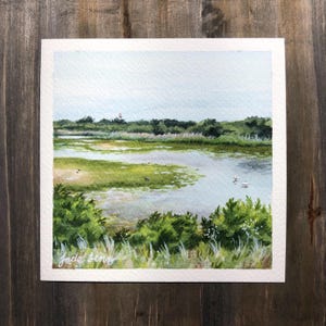 May include: Watercolor painting of a marsh landscape with a small body of water in the foreground. The water is a light blue color and there are green reeds and grasses growing in the water. There are a few white birds swimming in the water. The background is a light blue sky with a few clouds. The painting is signed "Jade Leong" in the bottom left corner.