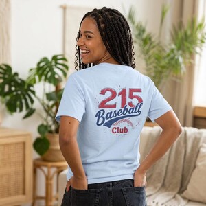 May include: A light blue t-shirt featuring a graphic on the back with the red text 215, the blue script text Baseball, and the red text Club, accompanied by a baseball illustration with red stitching and a distressed texture effect.
