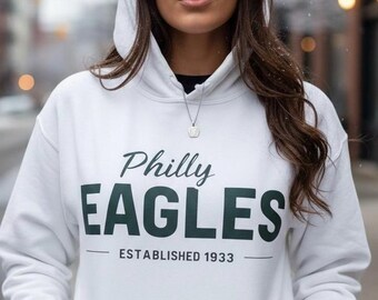 May include: White hoodie featuring the text "Philly EAGLES ESTABLISHED 1933" in dark green. The hoodie has a drawstring hood and a front pocket. The image is taken outdoors.