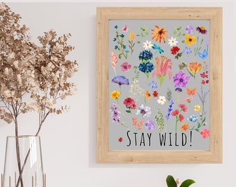 Può includere: Una stampa incorniciata con sfondo grigio che presenta un assortimento colorato di fiori ad acquerello e il testo "Stay Wild!" in lettere nere.