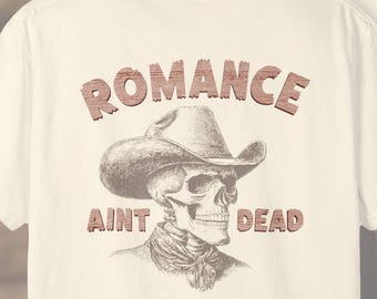 May include: Two off-white Comfort Colors t-shirts. One shirt features a skull wearing a cowboy hat with the text "ROMANCE AINT DEAD". The other shirt has a smaller skull graphic with a cowboy hat. The Comfort Colors logo is at the top.