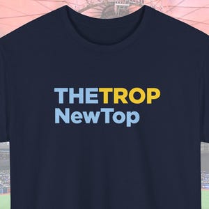 Può includere: T-shirt blu navy con la scritta "THETROP NewTop" in azzurro e giallo. La maglietta è mostrata in uno stadio, suggerendo un tema sportivo o di evento. Una firma è nell'angolo in basso a destra.