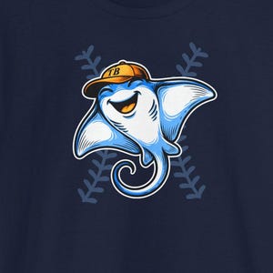 Pode incluir: Camiseta azul marinho com um desenho animado de arraia-manta usando um boné de beisebol com "TB". A arraia-manta está sorrindo, cercada por costuras de beisebol. A camisa está em um fundo de estádio de beisebol.