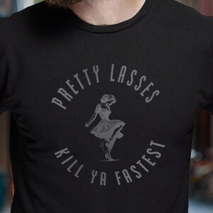Puede incluir: Camiseta negra con un gráfico circular. El texto "PRETTY LASSES KILL YA FASTEST" rodea una ilustración estilizada de una mujer con un vestido. La camiseta está hecha de un material suave.