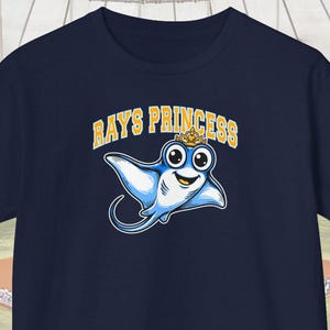 Pode incluir: Camiseta azul marinho com um desenho animado de arraia com uma coroa e o texto "RAYS PRINCESS" em dourado. A arraia é azul claro com um rosto sorridente. Uma peça de roupa casual.