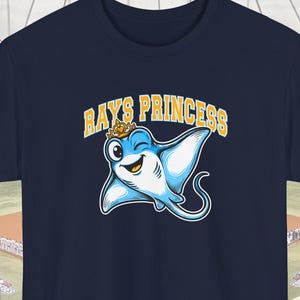 Pode incluir: Camiseta azul marinho com um desenho animado de arraia usando uma coroa e piscando. O texto "RAYS PRINCESS" está arqueado acima da arraia em amarelo. O fundo é um estádio de beisebol.