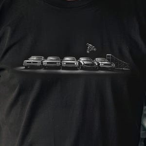 May include: A black short-sleeve t-shirt featuring a graphic print of five cars lined up side-by-side with a motorcycle jumping over them from a ramp. The design is centered on the chest area of the shirt, rendered in shades of white and gray.