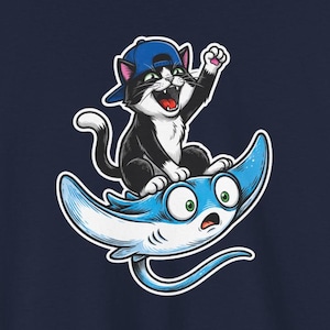 Puede incluir: Camiseta azul marino con un gato de dibujos animados con una gorra de béisbol azul, montando una manta raya azul. El gato saluda con una pata. La manta raya tiene una expresión de sorpresa. El fondo es un estadio de béisbol.