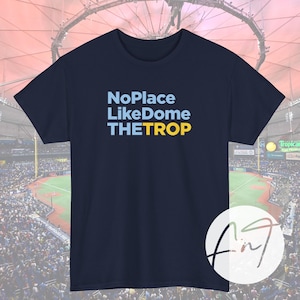 May include: Navy blue t-shirt with the text "No Place Like Dome THE TROP" in light blue and yellow. The shirt is a crew neck and appears to be made of cotton. The background shows a baseball stadium.
