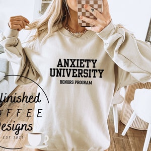 Könnte beinhalten: Ein beiger Pullover mit dem Aufdruck "Anxiety University Honors Program" in Schwarz. Der Pullover wird von einer Person mit braunen Haaren und einer braunen Mütze getragen.
