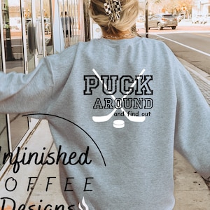 Pode incluir: Uma camisa de moletom cinza com o texto "Puck Around and find out" em preto com um gráfico de um taco de hóquei e um disco. A camisa de moletom está sendo usada por uma pessoa que caminha por uma calçada.
