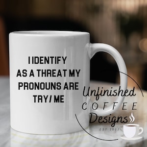 以下が含まれることがあります： 白いセラミック製のコーヒーカップに、黒い文字で「私は脅威として認識されています。私の代名詞は試してみてください/私です」と書かれています。カップは、大理石の表面に置かれています。背景には、「Unfinished Coffee Designs est 2023」という文字が見えます。