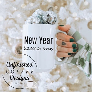 May include: White coffee mug with the text "New Year same me" and the logo "Unfinished Coffee Designs Est 2023" with a small coffee cup icon. The mug is filled with silver disco ball ornaments and is being held by a hand with green nail polish.