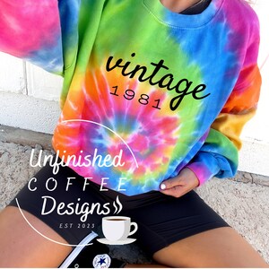 Pode incluir: Uma camisola tie-dye com o texto "vintage 1981" em preto. A camisola é de cor azul claro, verde, rosa e laranja. O texto "Unfinished Coffee Designs" está num círculo abaixo da camisola.