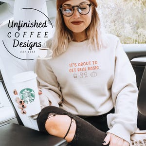 May include: A beige sweatshirt with the text "IT'S ABOUT TO GET REAL BASIC" in orange. The sweatshirt also features a coffee cup, bow, and pumpkin graphic. The image includes a coffee cup and the text "Unfinished Coffee Designs EST 2023".