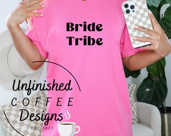 May include: Pink oversized t-shirt with the text "Bride Tribe" in black. The shirt has a small coffee cup graphic with the text "Unfinished Coffee Designs Est. 2023" on the bottom left corner.