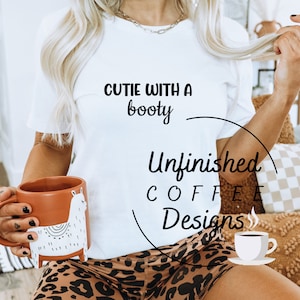 Peut inclure: Un t-shirt blanc avec le texte "Cutie with a booty" et le logo "Unfinished Coffee Designs". La personne portant le t-shirt tient une tasse brune avec un motif de lama blanc. La personne porte également un short à imprimé léopard.