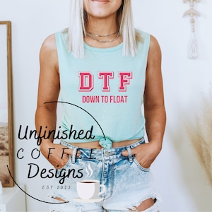 May include: A woman wearing a light blue sleeveless shirt with the text "DTF Down to Float" in pink and gray letters. She is wearing denim shorts and has her hands in her pockets.