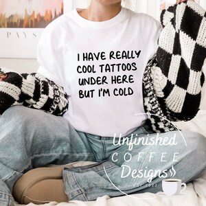以下が含まれることがあります： 黒文字で「I have really cool tattoos under here but I'm cold」とプリントされた白いTシャツ。ベッドに座って黒と白のチェック柄の毛布をかけている人が着ています。