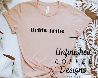 May include: A peach-colored t-shirt with the text "Bride Tribe" in black. The shirt is folded and tied in a knot. The background is white with a brown and white floral pattern.