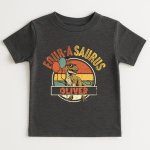 Detailed view of the nameplate customization on a dinosaur birthday shirt. The name "OLIVER" is written in a classic block font inside a vintage-style border. Above the name, the cool T-Rex with sunglasses signifies the "Four-a-Saurus" milestone. The distressed finish on the graphic provides a trendy, lived-in look for modern kids.